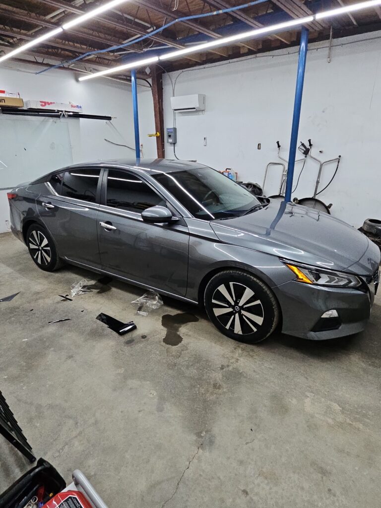 T'S Window Tinting Applying Window Film To A Grey Sedan In A Garage Setting With Tools And Equipment Nearby