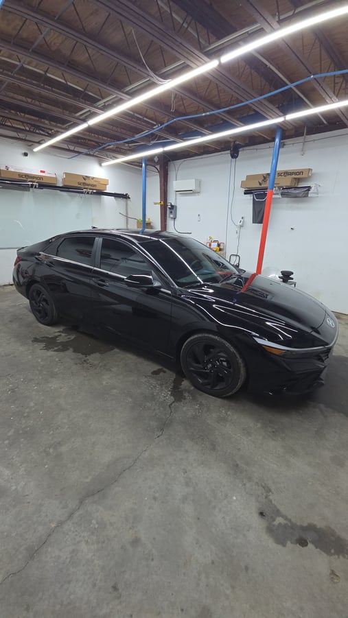 A sleek black Hyundai Elantra sits in the T’s Window Tinting shop with freshly tinted windows and matching black wheels