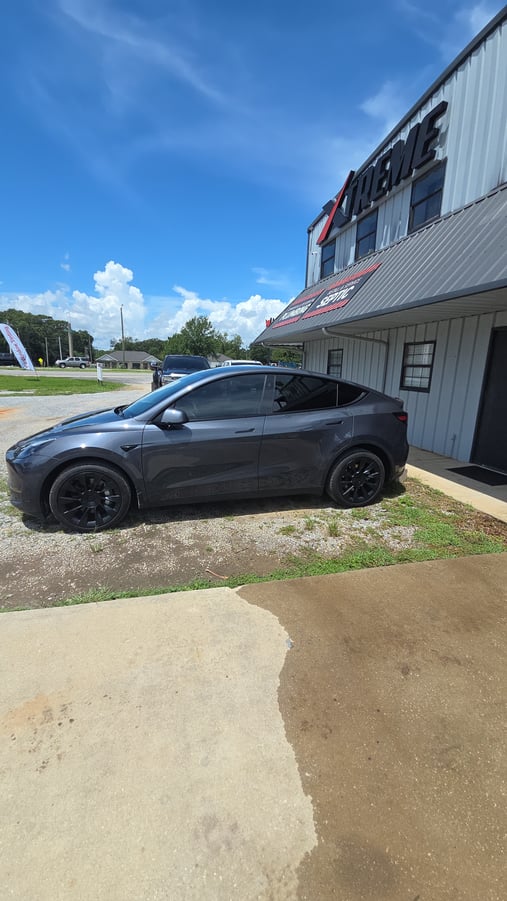 A dark gray Tesla Model Y with freshly tinted windows is parked outside the shop on a sunny day