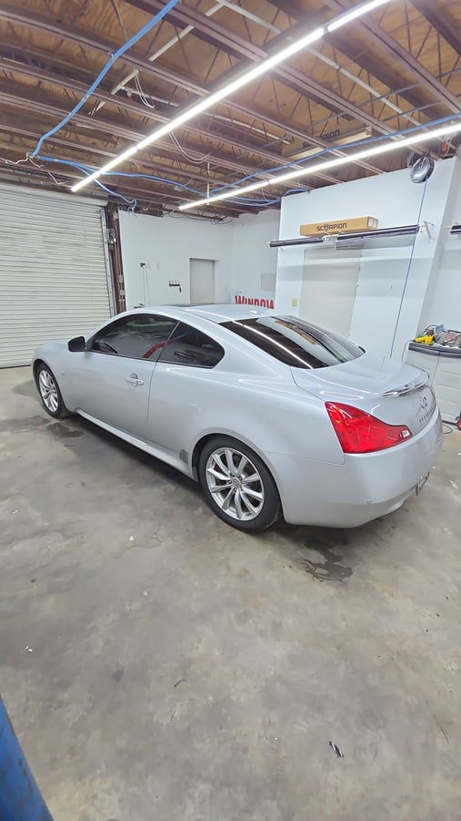 A silver Infiniti Q60 coupe sits inside the T’s Window Tinting shop with freshly tinted side and rear windows